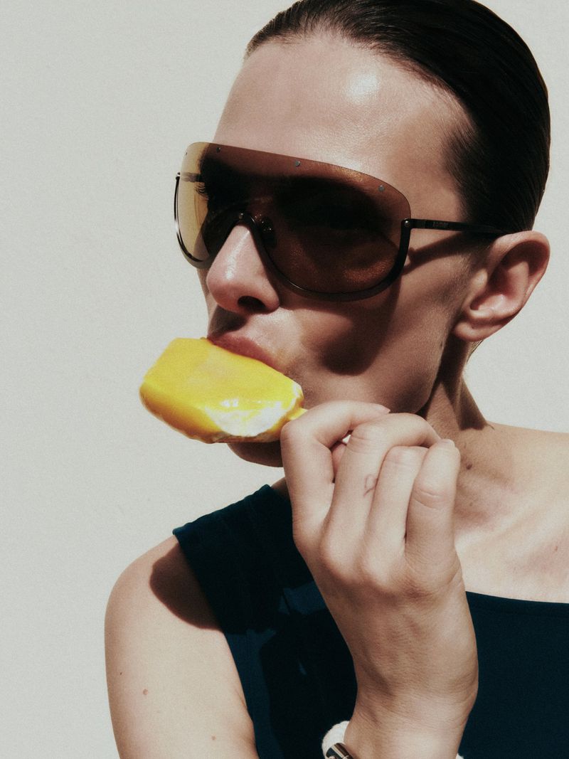 Model Tanya Ruban wearing oversized shield sunglasses and eating a yellow popsicle in direct sunlight against a pale backdrop.
