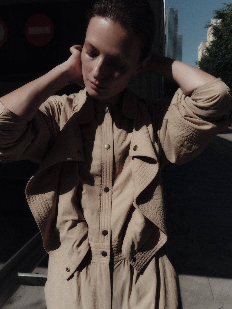Model Tanya Ruban wearing a tan button-down jumpsuit with hands behind her head in an outdoor urban setting for Duel Magazine.