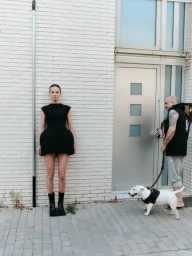 Model Tanya Ruban in a structured black mini dress standing against a white brick wall as a man walks a white bull terrier past a door.