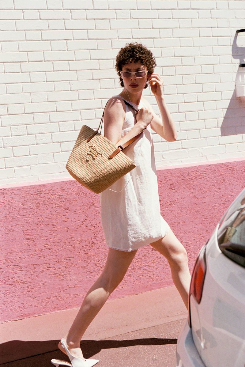 Model Jesi Evans walks along a street wearing a white dress, sunglasses, and a woven tote bag next to a pink painted brick wall.