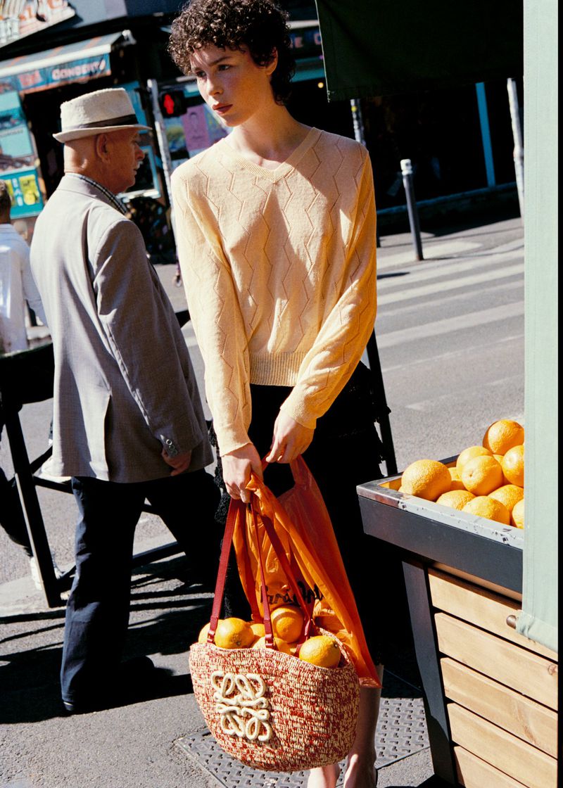 Model carrying a woven basket tote filled with oranges on a sunny city sidewalk next to a fruit stand.