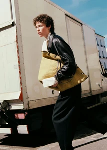 Model walking on a city street in a black leather jacket carrying an oversized yellow woven tote bag beside a white truck.