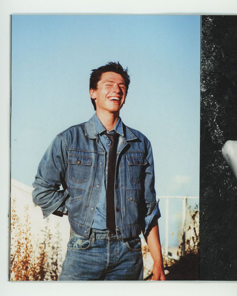 A smiling man wearing a denim jacket and tie outdoors against a clear blue sky background.