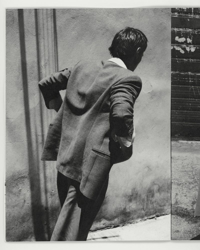 Man in a tailored wool blazer seen from behind, walking alongside a sunlit stucco wall with linear shadows.