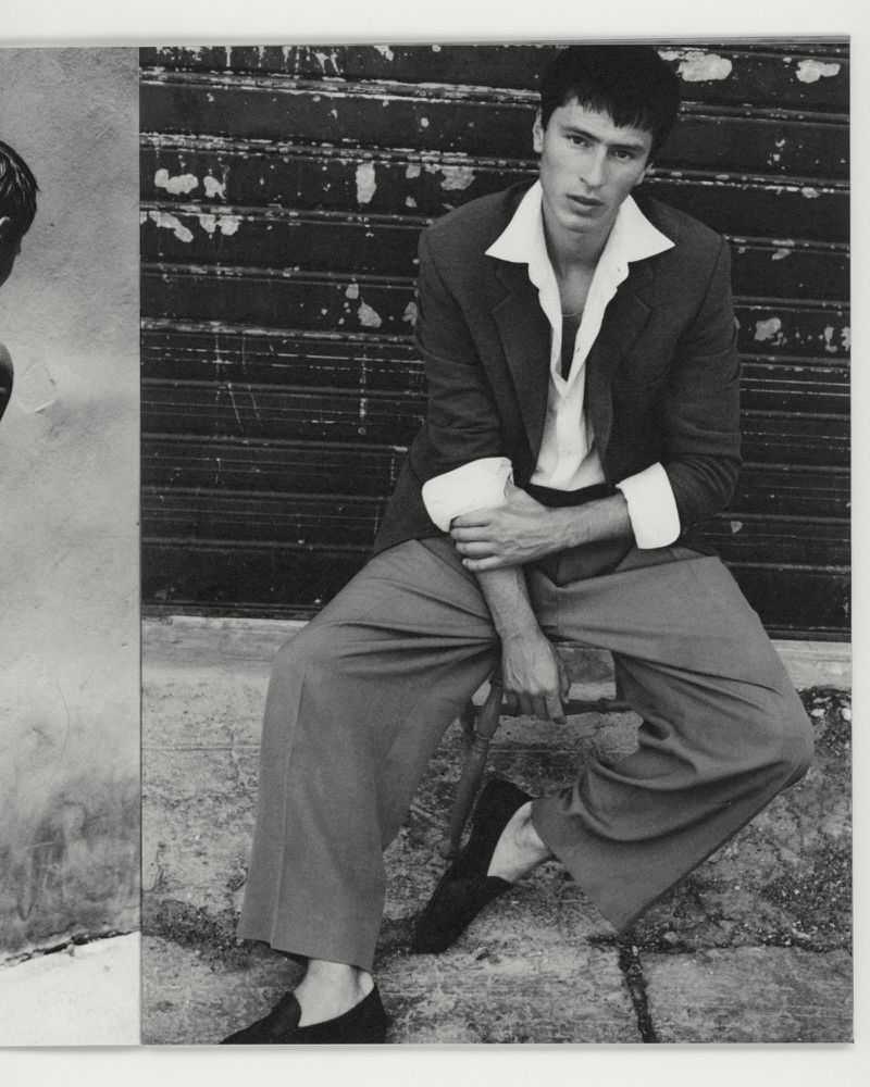 Black and white portrait of a man seated wearing a tailored jacket and wide trousers against a slatted metal shutter background