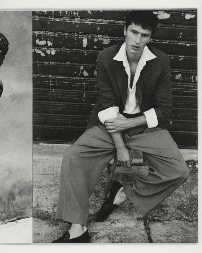Black and white portrait of a man seated wearing a tailored jacket and wide trousers against a slatted metal shutter background