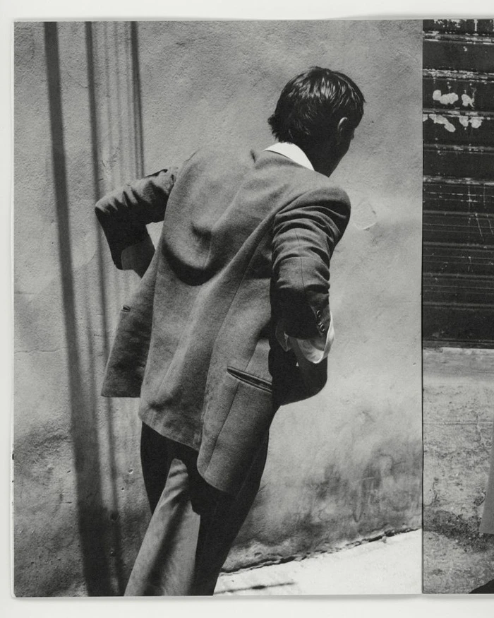 Man in a tailored wool blazer seen from behind, walking alongside a sunlit stucco wall with linear shadows.