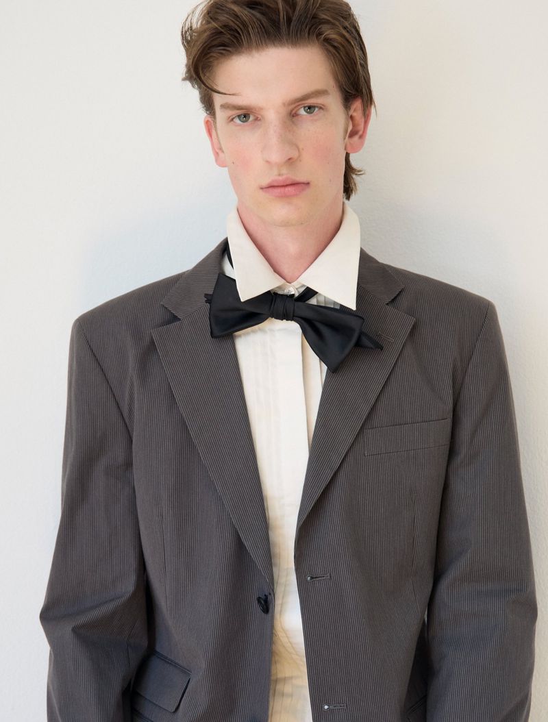 Portrait of a young man wearing a grey suit jacket, white collared shirt, and black bow tie against a white background.
