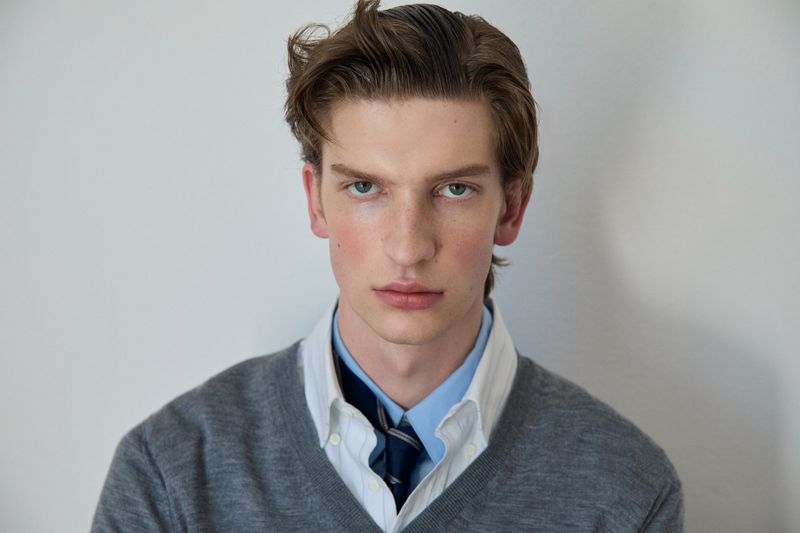 Portrait of a young man wearing a grey v-neck sweater over layered collared shirts with a tie against a plain studio backdrop.