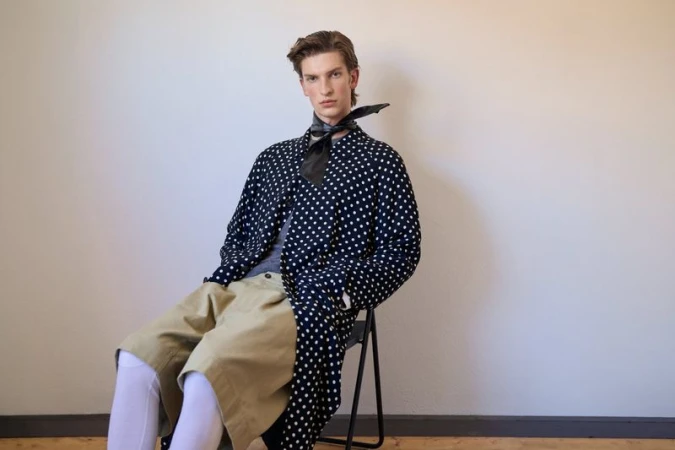 A young man seated on a chair wearing a navy polka dot coat, beige shorts, and white tights against a plain wall.