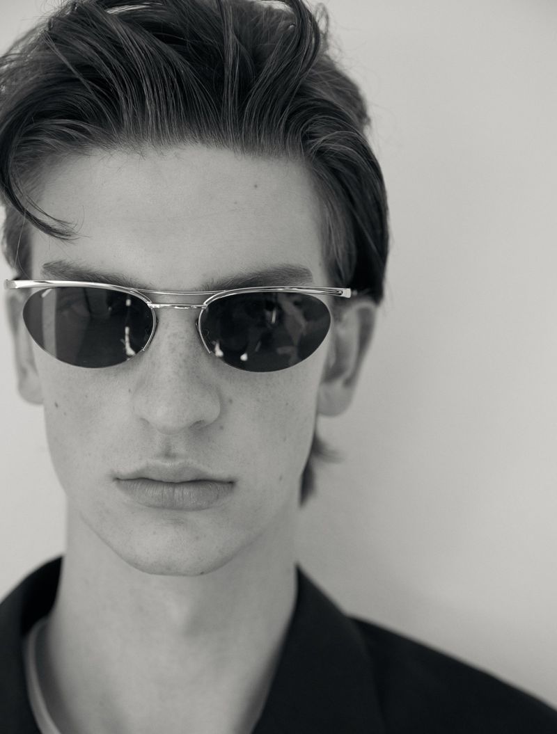 Close-up black and white portrait of a young man wearing metallic oval sunglasses and a dark shirt against a pale background.