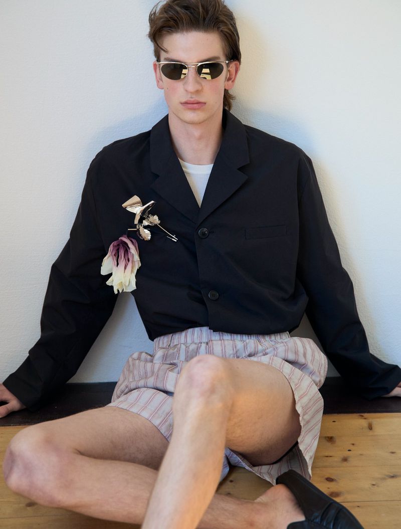 A young man in silver sunglasses, a black jacket pinned with a flower, and striped shorts sits against a white wall.
