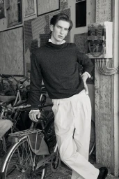 Model in a dark knit sweater and white pleated trousers posing with a bicycle against a textured wall.
