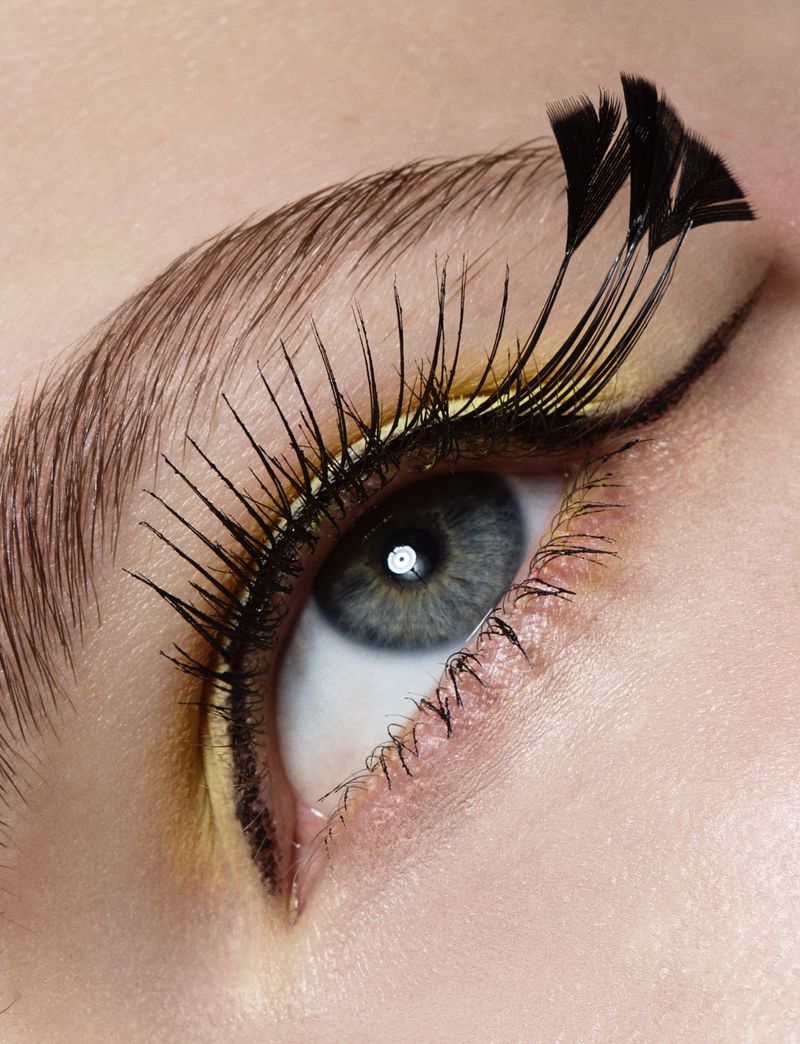 Extreme close-up of a blue eye featuring precise graphic eyeliner, pale yellow liner, and dramatic black feathered false eyelashes.
