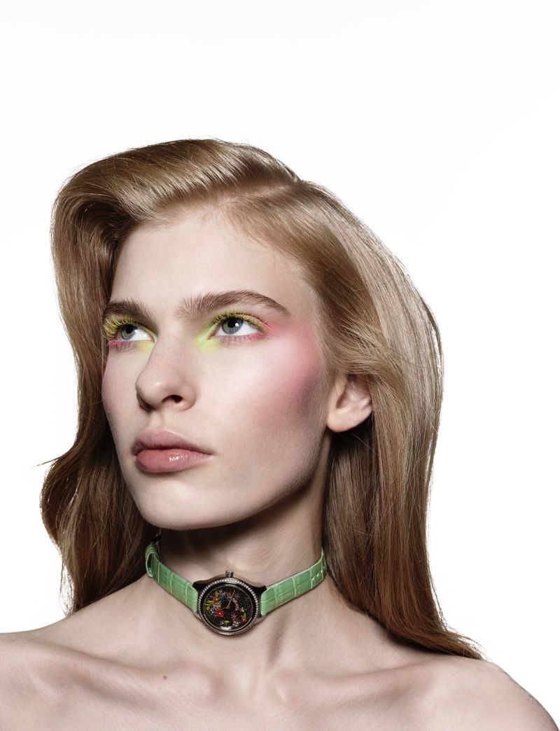 Close-up portrait of a model with pastel eye makeup wearing a green watch around her neck against a white background.