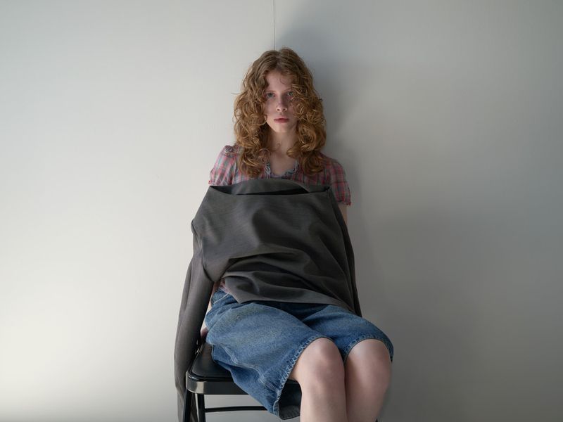 Woman with curly red hair sits indoors, wearing a plaid shirt and denim shorts, with a blazer draped over her lap against a white wall.