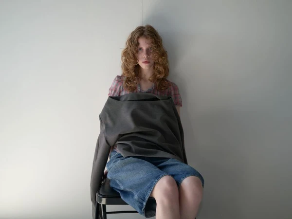 Woman with curly red hair sits indoors, wearing a plaid shirt and denim shorts, with a blazer draped over her lap against a white wall.