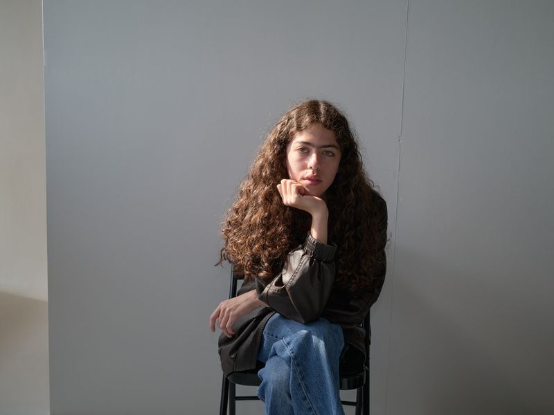 A young woman with long, curly brown hair is sitting in a chair in front of a grey wall, looking directly at the camera.