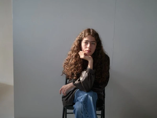 A woman with long curly hair resting her chin on her hand while seated on a black chair wearing a dark jacket and blue jeans.