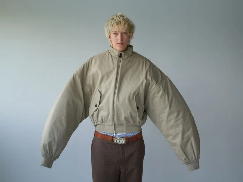 Person in an oversized, neutral-tone jacket, brown pants, and a statement belt stands in a studio with a pale gray background.