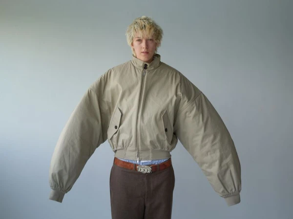 Person in an oversized, neutral-tone jacket, brown pants, and a statement belt stands in a studio with a pale gray background.