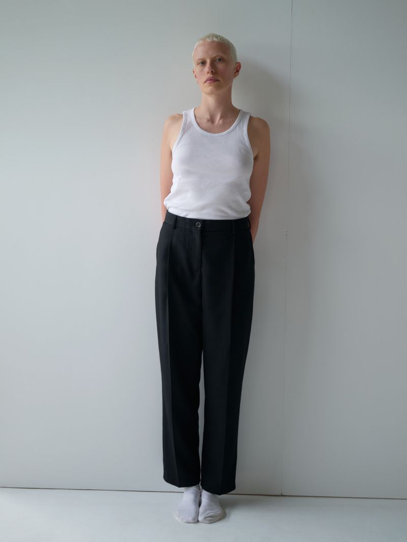 A model with a blonde buzz cut standing against a plain white wall wearing a white tank top, black tailored trousers, and white socks.