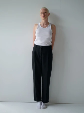 A model with a blonde buzz cut standing against a plain white wall wearing a white tank top, black tailored trousers, and white socks.