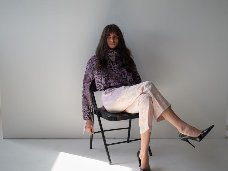 Adult female with dark hair, wearing a purple animal-print blouse, patterned pants, and black high heels, seated on a black folding chair.