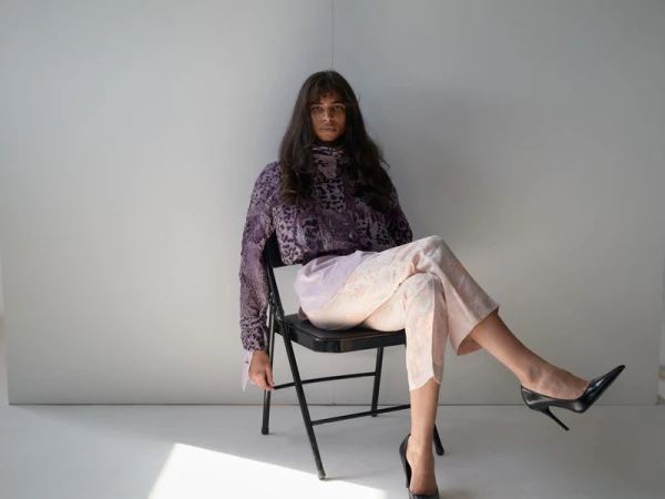 Adult female with dark hair, wearing a purple animal-print blouse, patterned pants, and black high heels, seated on a black folding chair.