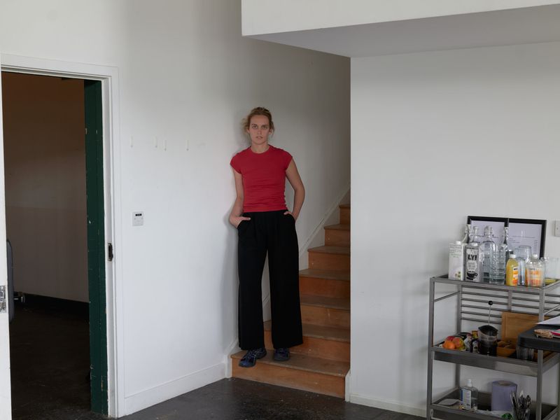 Woman in red shirt and black pants poses on wooden stairs in a bright white interior with a metal cart and open doorway.