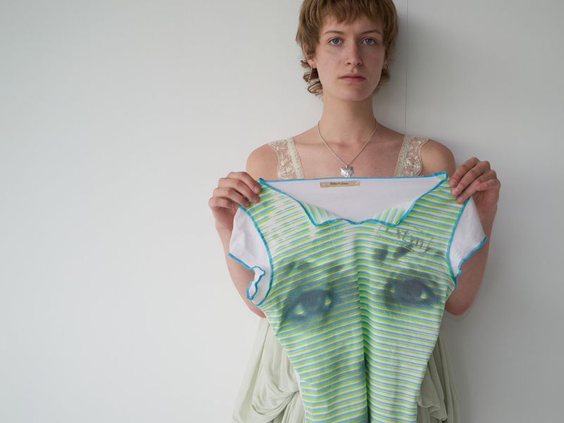 Woman holds a green and white striped shirt with a faded image of eyes in front of a plain white wall with natural lighting.