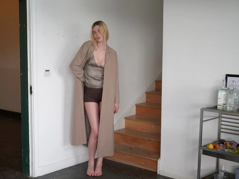 A young fair-skinned woman stands indoors wearing a long beige coat, a shimmery green top, and brown shorts near wooden stairs.