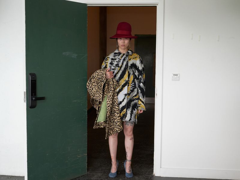 Woman in patterned coat and hat stands in doorway holding leopard print garment, casual fashion against a white wall with a dark door.