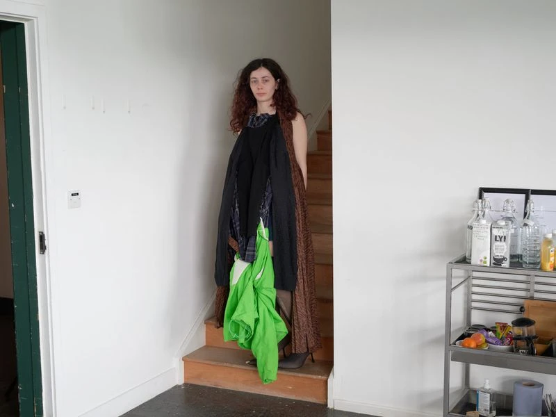 A woman stands indoors on stairs with clothing draped on her, next to white walls and a metal cart with bottles and glassware.