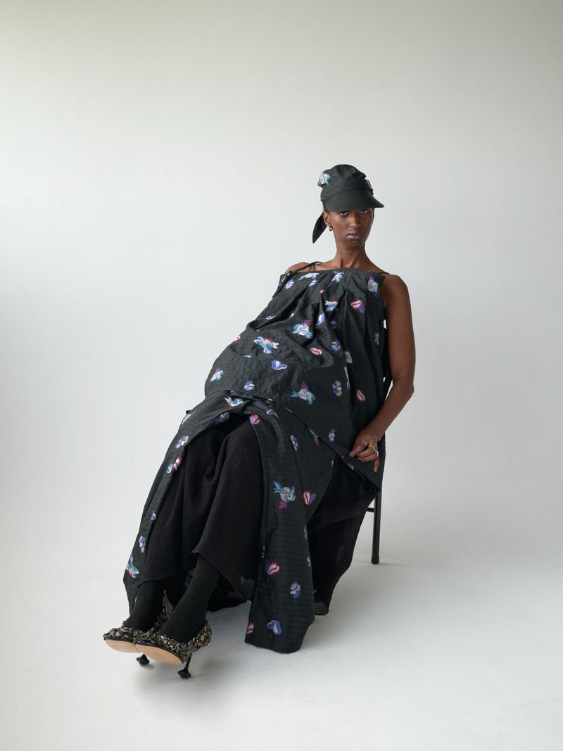 An adult female wearing a black patterned dress and hat sitting on a chair in a photo studio against a plain white background.