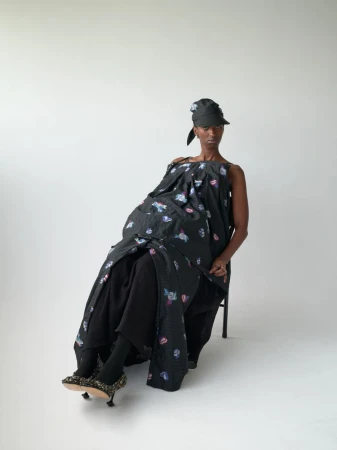 An adult female wearing a black patterned dress and hat sitting on a chair in a photo studio against a plain white background.