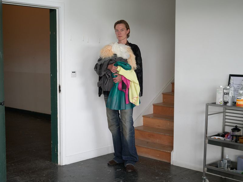 A person standing indoors holding a large pile of colorful clothes, with a glimpse of a staircase and trolley in a brightly lit room.