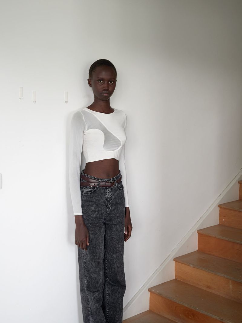 Full-length portrait of a slim, dark-skinned woman in a long-sleeved white top, acid-wash jeans, and a chain belt standing on a staircase.