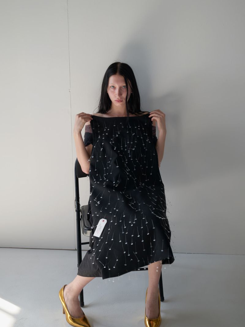 A fair-skinned woman with long dark hair poses in a photo studio, sitting on a black chair while holding a black dress with pearl embellishments.