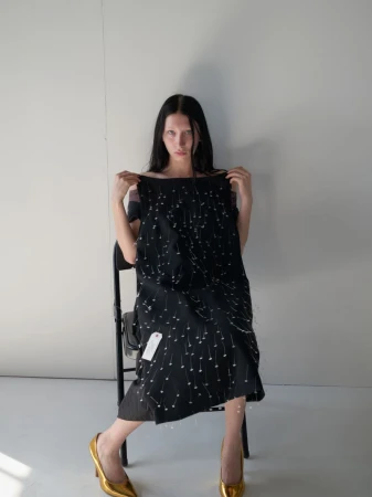 A fair-skinned woman with long dark hair poses in a photo studio, sitting on a black chair while holding a black dress with pearl embellishments.