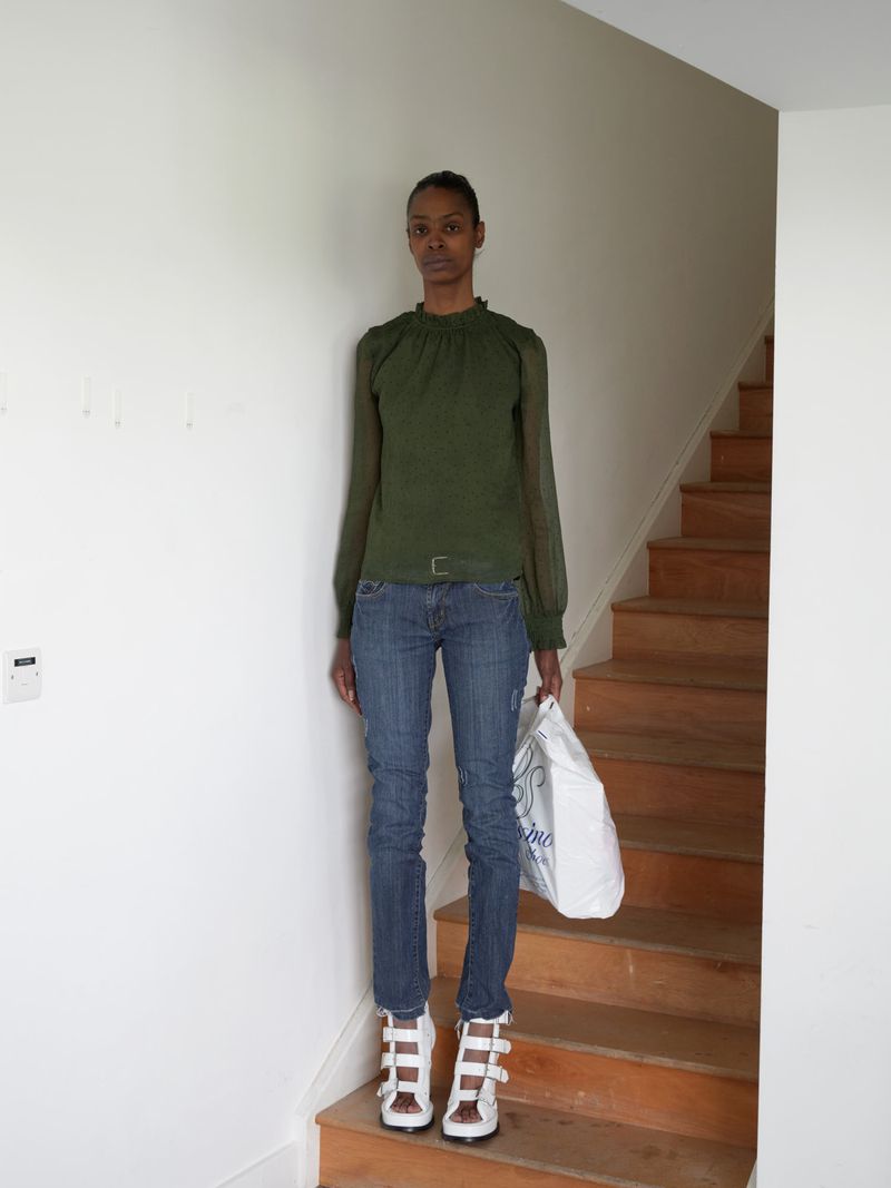 Full-length portrait of an adult female standing on a wooden staircase wearing a green top, blue jeans, and white high-heeled sandals.