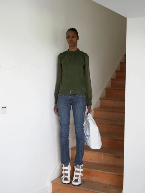 Full-length portrait of an adult female standing on a wooden staircase wearing a green top, blue jeans, and white high-heeled sandals.