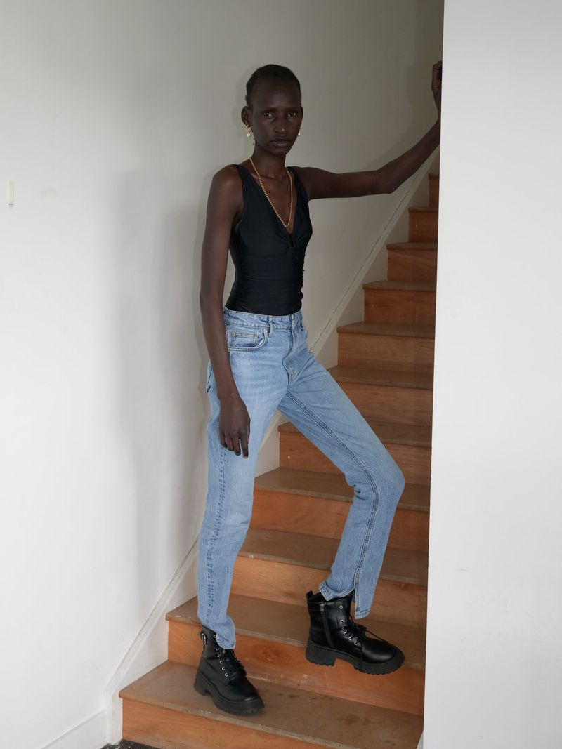 A slender woman in a black top, light blue jeans, and black boots poses on wooden stairs against a white wall indoors.