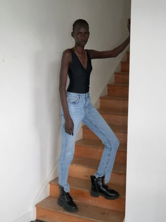 A slender woman in a black top, light blue jeans, and black boots poses on wooden stairs against a white wall indoors.