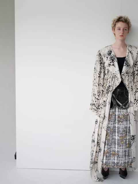 Woman in a patterned, flowing coat and patterned skirt stands in a white photo studio, wearing dark shoes and a black top.