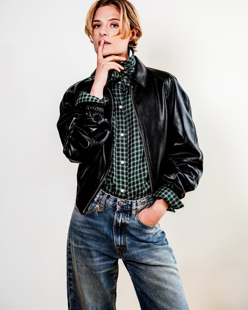 Actor Louisa Jacobson wearing a black leather jacket, green plaid shirt, and blue denim jeans against a white backdrop.