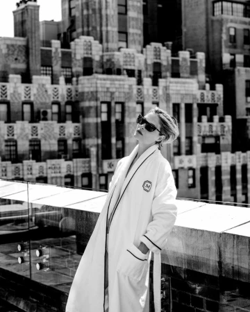 Louisa Jacobson wearing dark sunglasses and a white piped bathrobe on a rooftop overlooking city skyscrapers in black and white.
