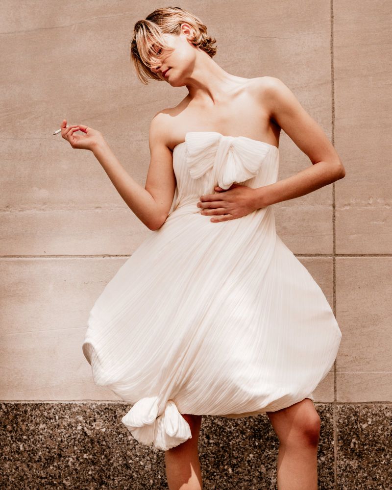 Louisa Jacobson wearing a strapless white pleated bubble dress while posing outdoors against a stone wall.