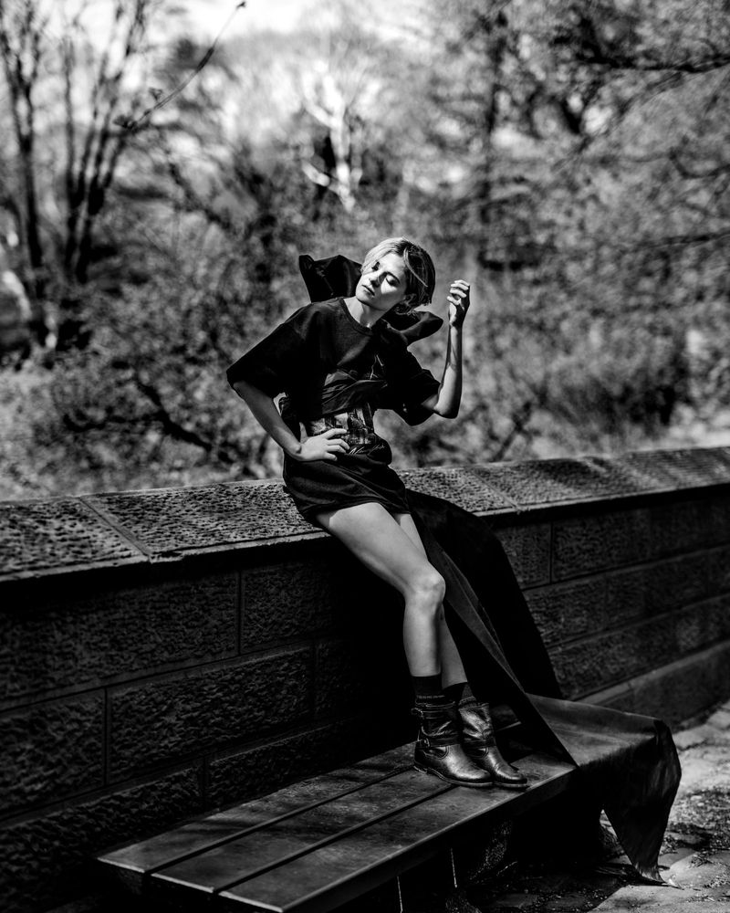 Louisa Jacobson wearing a sculptural black mini dress and boots, seated against a stone wall in an outdoor park in black and white.