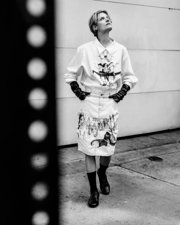 Black and white portrait of Louisa Jacobson in a graphic white shirt, printed skirt, and black gloves standing outdoors.